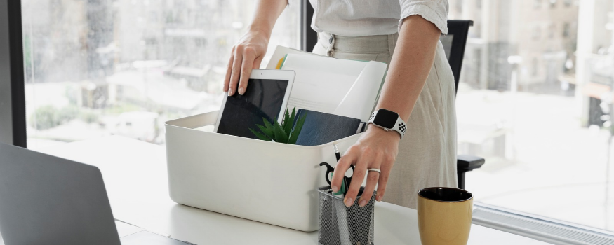 Freistellung nach Kündigung: Mitarbeiterin räumt ihren Arbeitsplatz und packt Laptop, Unterlagen und Büromaterial in eine Kiste.