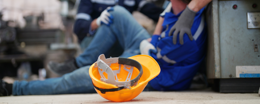 Ein Bauarbeiter sitzt nach einem Arbeitsunfall verletzt auf dem Boden, während ein Kollege ihm hilft. Im Vordergrund liegt ein gelber Schutzhelm – Symbol für Arbeitssicherheit und die Bedeutung der Unfallversicherung durch die Berufsgenossenschaft.