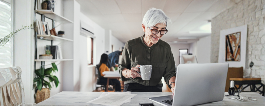 Ältere Frau arbeitet lächelnd am Laptop in einem modernen Büro und hält eine Kaffeetasse. Das Bild steht für die Aktivrente und zeigt erfahrene Beschäftigte, die auch nach Erreichen der Regelaltersgrenze weiterarbeiten.