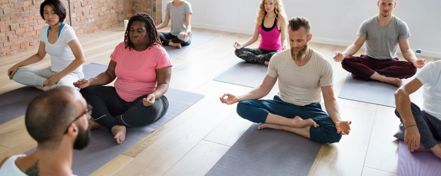 Gruppe von Mitarbeitenden sitzt in einem Yoga-Kurs im Schneidersitz und meditiert – Beispiel für betriebliche Gesundheitsförderung, die Arbeitgeber steuerfrei anbieten können.