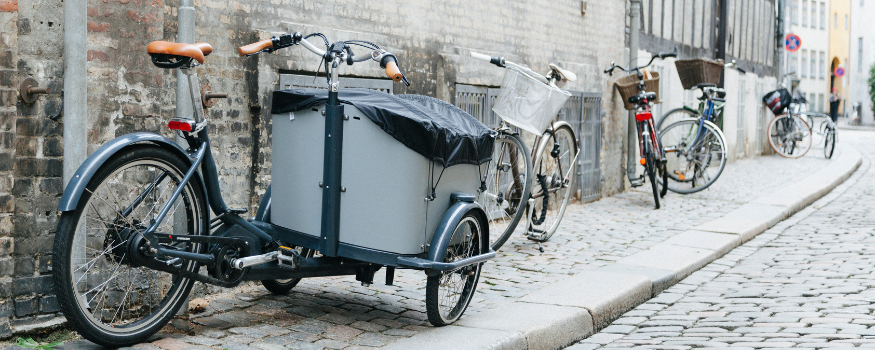 Straßenszene mit mehreren Fahrrädern, im Vordergrund ein betriebliches E-Lastenrad mit großer Transportbox – ideal für Unternehmen und Selbstständige, die Waren oder Einkäufe klimafreundlich und kostengünstig transportieren möchten.