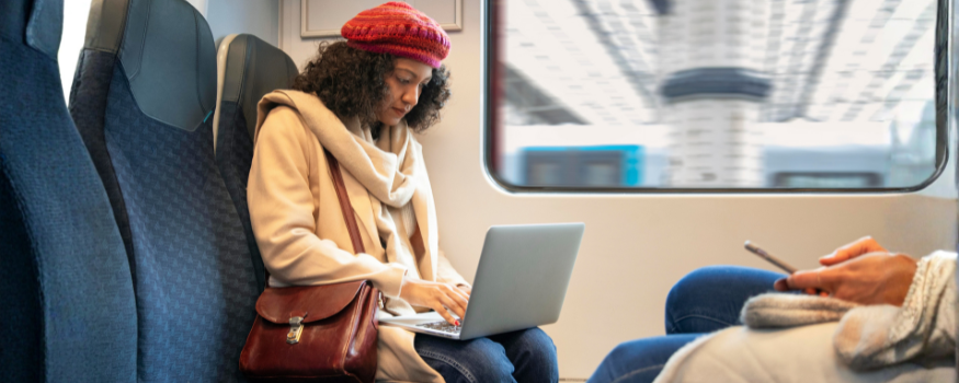 Junge Frau sitzt im Zug am Fenster und arbeitet mit einem Laptop auf dem Schoß. Sie trägt Mütze und Mantel, neben ihr liegt eine Tasche. Das Bild steht für Pendeln im öffentlichen Nahverkehr – etwa mit dem Deutschlandticket als Jobticket vom Arbeitgeber.