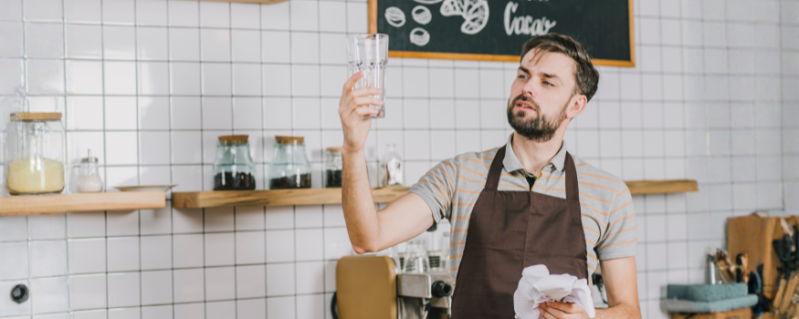 Ein Mann mit brauner Schürze steht in einem Café und prüft ein frisch poliertes Glas gegen das Licht. Hinter ihm sind weiße Fliesen, Küchenutensilien und Regale mit Vorratsgläsern zu sehen – typisch für einen kleinen Gastronomiebetrieb mit Minijobbern.