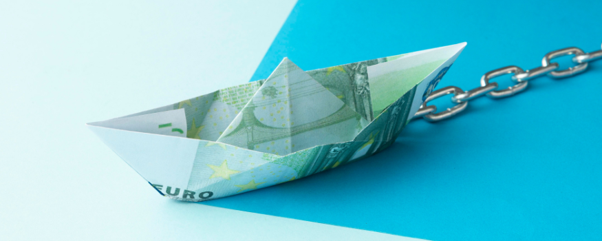 Gefaltetes Papierboot aus einem Euro-Schein, das an einer Kette befestigt ist – Symbol für gebundenes Vermögen und eingeschränkte Verfügbarkeit von Kapital, passend zur Idee der GmgV und ihrer Vermögensbindung.