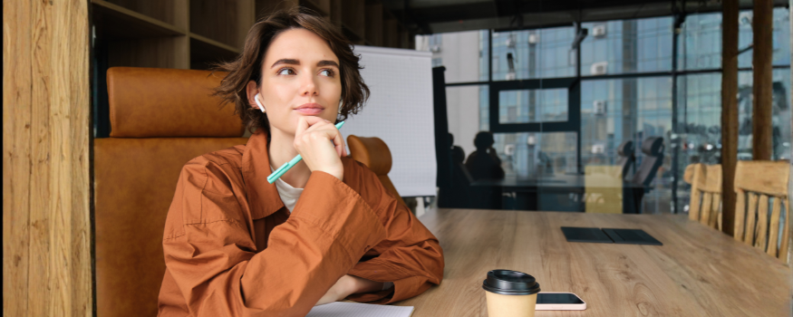 Eine Frau sitzt nachdenklich im modernen Büro, Stift und Notizbuch vor sich. Das Bild symbolisiert die Überlegung, ob eine GmbH oder eine UG die passende Rechtsform für das eigene Unternehmen ist.