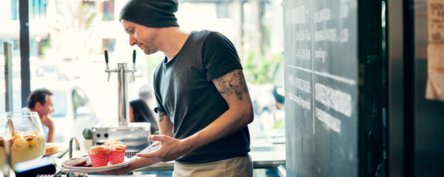 Ein Café-Mitarbeiter mit Tattoos und Mütze serviert Muffins und Kuchen. Das Bild symbolisiert typische Beschäftigungen in Gastronomie und Service und eignet sich zur Veranschaulichung von Themen wie Midijob vs. Minijob.