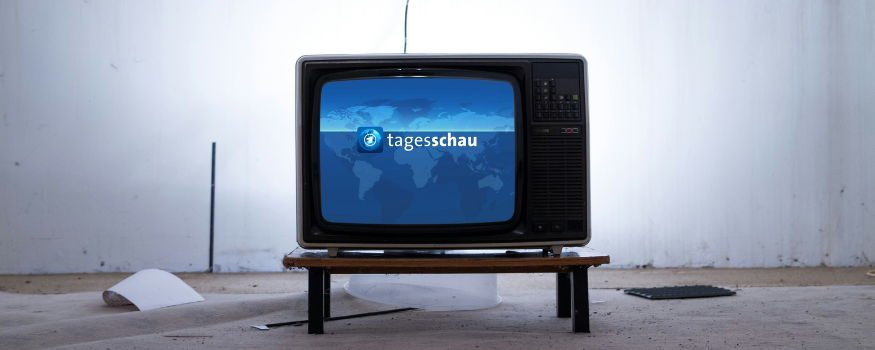 Ein alter Röhrenfernseher mit dem Logo der „Tagesschau“ auf dem Bildschirm steht auf einem niedrigen Holztisch in einem leeren Raum.