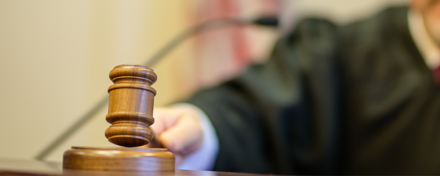 Richter schlägt mit einem hölzernen Richterhammer auf das Pult. Der Fokus liegt auf dem Hammer im Vordergrund, die Robe der Justizperson ist unscharf im Hintergrund erkennbar.