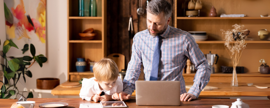 Ein Vater arbeitet im Homeoffice am Laptop, während sein kleines Kind neben ihm am Küchentisch ein Tablet benutzt. Das Bild zeigt die Vereinbarkeit von Beruf und Familie und verdeutlicht, wie wichtig familienfreundliche Arbeitsmodelle für Eltern sind.